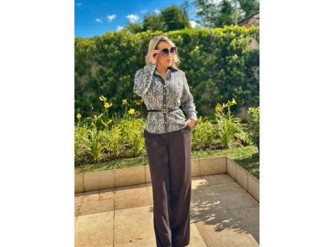 Mulher usando camisa social feminina com estampa, calça marrom de alfaiataria e óculos de sol, em um look elegante ao ar livre.