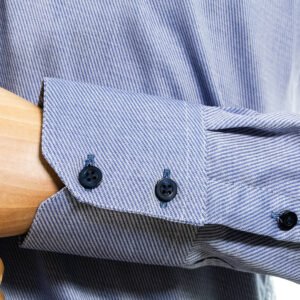 Detalhe do punho da camisa azul em algodão, com botões escuros alinhados e acabamento de alfaiataria na barra do punho.