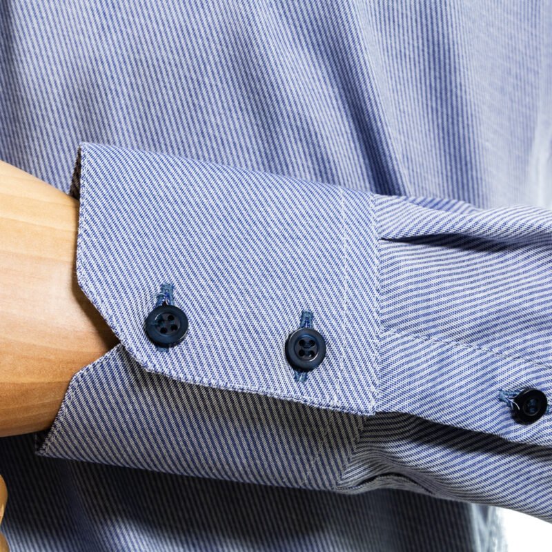 Detalhe do punho da camisa azul em algodão, com botões escuros alinhados e acabamento de alfaiataria na barra do punho.