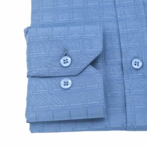 Close do punho com botão da camisa social azul com padronagem xadrez larga, reforçando o acabamento elegante.